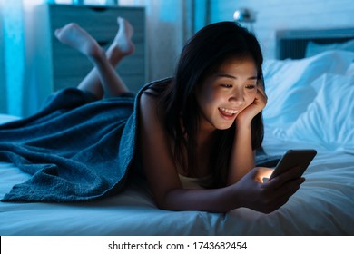 Happy Asian Japanese Teen Lying In Front In Bed At Home With Smartphone. Young Joyful Woman Watching Funny Video On Mobile Phone In Bedroom At Night And Laughing. Relax Girl Shopping Online Indoors