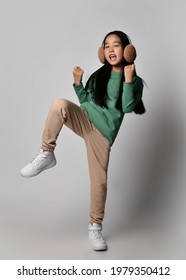 Happy Asian Kid Girl In Green Earmuffs, Sportswear And White Sneakers Stands On One Leg Holding Knee Up And Gesturing, Singing, Screaming Over Gray Background