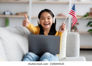 Happy Asian Kid Pretty Long-haired Girl Learning English On Internet, Sitting On Couch In Her Room, Holding Flag Of The US And Showing Thumb Up, Using Wireless Headset And Modern Laptop