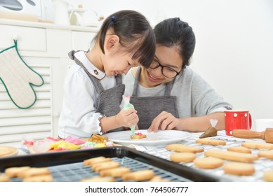 Happy Asian Kid And Young Mother Decorating Cookies In The Kitchen.