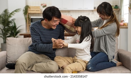 Happy Asian Korean Father Mother And Daughter Playing Tickle On Living Room Couch At Home. The Woman Having Fun Hitting The Man With Cushion