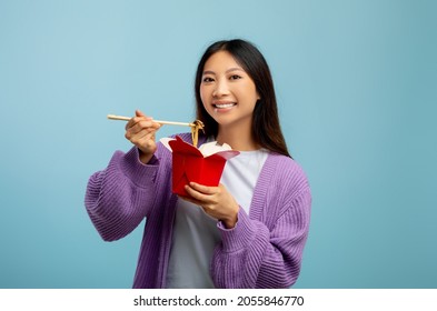 Happy Asian Lady Eating Noodles With Chopsticks, Holding Paper Box And Smiling To Camera, Standing On Blue Studio Background. Food Delivery, Fastfood Concept