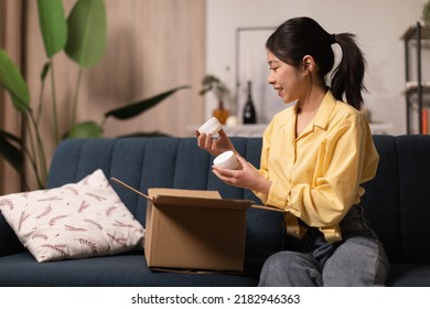 Happy Asian Lady Holding Delivered Moisturizer Jars Unpacking Cardboard Box After Successful Beauty Shopping Sitting On Sofa At Home. Cosmetology Products, Delivery Service Concept