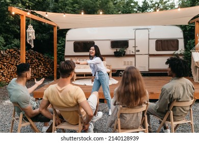 Happy Asian lady playing word guessing game with diverse friends near motorhome at campsite. Group of young people enjoying charades or pantomime riddles. Fun outdoor entertainments