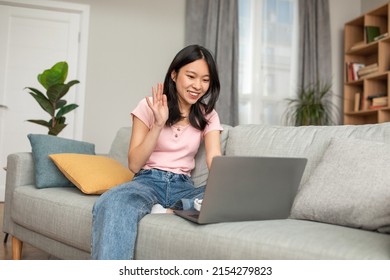 Happy Asian Lady Video Calling Via Laptop Computer, Waving Hello At Webcam, Sitting On Sofa In Living Room. Cheerful Korean Woman Communicating Online, Using Modern Gadget At Home