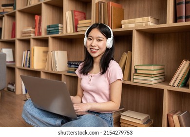 Happy Asian Lady In Wireless Headphones Typing On Laptop Keyboard While Leaning On Bookshelf And Smiling At Camera. Young Woman Working Or Study Online On Computer