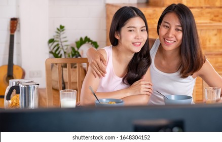 Happy Asian Lesbian Couple Watching Television While Eating Breakfast At Kitchent In Cozy Home