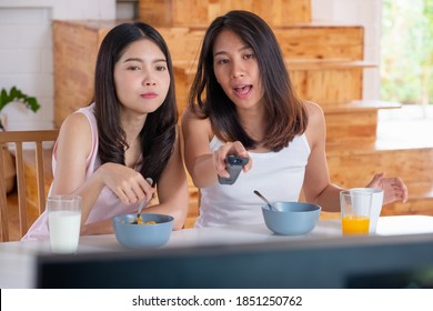 Happy Asian Lesbian Couple Watching Television While Eating Breakfast At Kitchent In Cozy Home