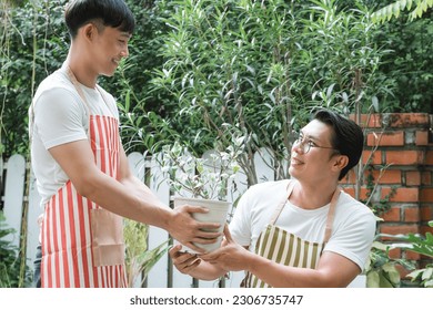 Happy Asian LGBTQ gay men couple wear apron shoveling watering a plant pot outdoors at home. Young man smiling cheerfully, giving a pot plant to boyfriend. Lgbtq lifestyle, love, valentine day concept - Powered by Shutterstock