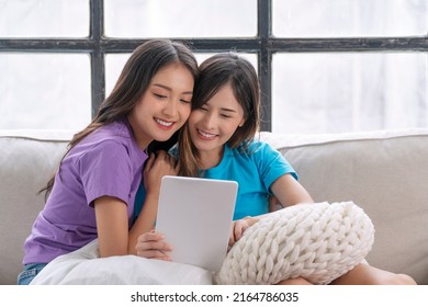 Happy Asian LGBTQ Lesbian Woman Looking Tablet Sitting Together Pillows On Sofa Living Room. Lover Young Couple Girl Use Tablet Purchase Shopping Online Sitting Relaxed On Couch At Home. Technology