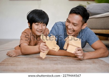 Happy Asian little boy playing a wooden figure model with his dad while laying on the living room floor together.