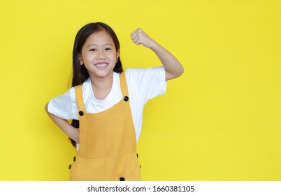 Happy asian little child girl showing his muscle with looking camera isolated on yellow background with copy space. Strong kid in dungarees with good health concept