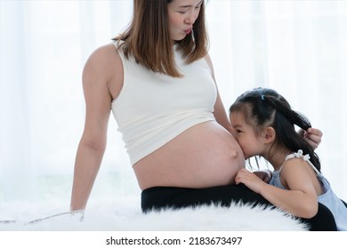 Happy Asian Little Cute Kid Girl Playing With Pregnant Mom On Bed At Home, Touch And Kiss Mother Belly With Tender And Love. Family Bond Concept. White Background