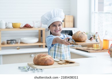 Happy Asian Little Cute Kid Girl Wear Chef Hat And Apron Holding Showing Homemade Whole Grains Bread On Wooden Tray In Kitchen At Home Or Cooking Class At School. Child Education Concept