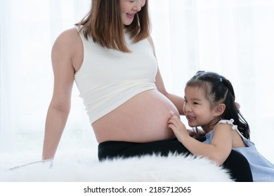 Happy Asian Little Cute Kid Girl Playing With Pregnant Mom On Bed At Home, Touch Mother Belly And Navel With Tender And Love. Family Bond Concept. White Background