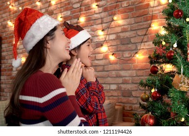 Happy Asian Little Girl And Mom Finally Finished Decorating Christmas Tree And Joyfully Enjoy Looking At The Done Work. Merry Xmas And Happy New Year Are Coming. Beautiful Small Family At Bright Home