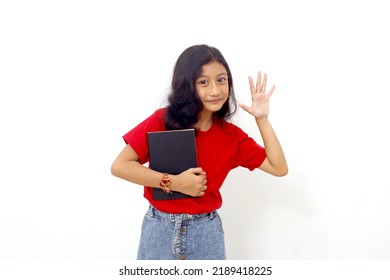 Happy asian little girl standing while showing five fingers and holding a book. Isolated on white