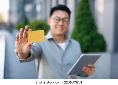 Happy Asian Male Entrepreneur Holding Tablet And Showing Credit Card To Camera, Selective Focus, Closeup. Man Shopping On Internet While Having Break Outdoors