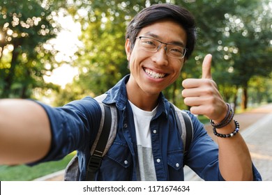 Happy Asian Male Student In Eyeglasses Making Selfie And Showing Thumb Up While Looking At The Camera Outdoors