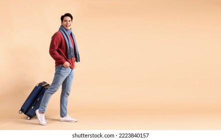 Happy Asian male tourist with luggage in red sweater and scarf enjoying winter vacation isolated on beige color background. Excited fashionable 20s guy going on a trip - Full body length studio shot. - Shutterstock ID 2223840157