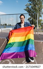 Happy Asian Man In Eyeglasses Sitting On Ramp In Skate Park With Lgbt Flag