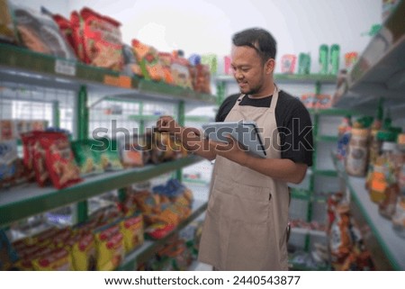 Happy Asian man as groceries or modren market staff checking products and goods on the display shelf while holding tablet and wearing grey apron