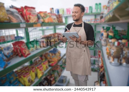 Happy Asian man as groceries or modren market staff checking products and goods on the display shelf while holding tablet and wearing grey apron
