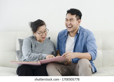 Happy Asian Man And His Mother Looking At Photo Album Together While Staying At Home.