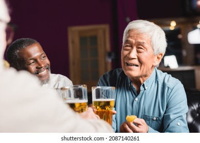 Happy Asian Man Holding Beer And Chips Near Blurred Multiethnic Friends In Pub