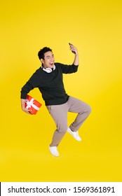 Happy Asian Man Holding Gift Box And Smart Phone, Jumping On Yellow Background.