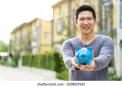 Happy Asian Man Holding Piggy Bank With Front Outdoor Home Background Copy Space On Left.Smiling Rich Property Owner Loan, Mortgage Or Installment For House.Investment In Real Estate Business And Debt