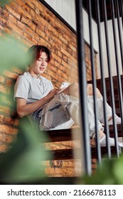 Happy Asian Man Looking At Mobile Phone Screen And Sitting On Stairs