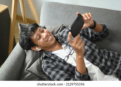 Happy Asian Man Lying On Couch And Watching Funny Video On Smart Phone.