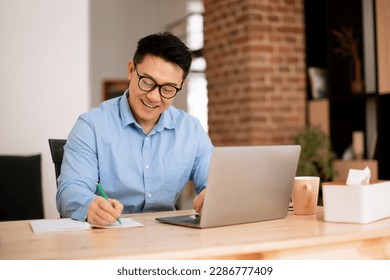 Happy asian man making notes, analyzing data for investment at workplace with laptop computer in home office interior. Work, business and startup planning, brainstorming with device remotely - Powered by Shutterstock