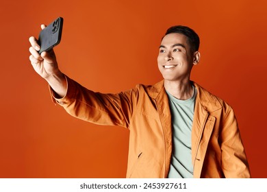 happy Asian man in stylish attire taking a selfie with his cell phone against an orange studio background. - Powered by Shutterstock