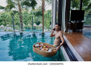 Happy Asian Man In Swimsuit Having Floating Breakfast Tray In Luxury Pool Hotel While Talking On The Phone. Young Male Enjoying Morning Coffee In Tropical Resort And Working Remotely On Online Project