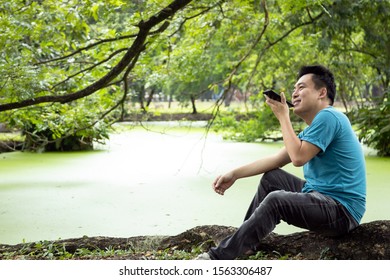 Happy Asian Man Talking On Smart Phone With The Digital Voice Assistant, Sitting In Green Nature, Male People Using Voice Command Recorder Or Calling On Speakerphone With Family, Technology Concept