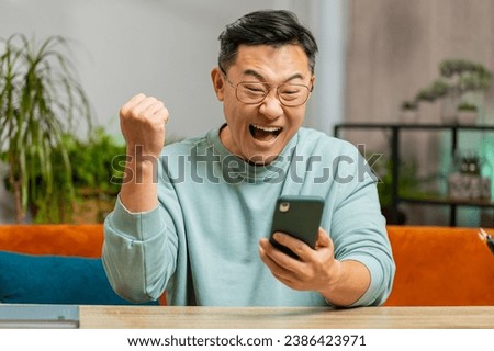 Happy Asian man use mobile smartphone typing browsing say wow yes celebrating success victory winning lottery jackpot goal achievement play game good positive news triumph. Guy at home in room on sofa
