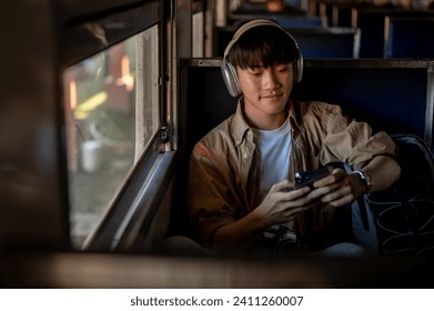 A happy Asian man is using his phone and listening to music on his headphones while he is on a train to somewhere. commuting, traveler, backpacker, solo traveler - Powered by Shutterstock