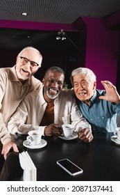 Happy Asian Man Waving Hand Near Senior Multiethnic Friends In Cafe