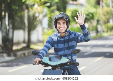 Happy Asian Man Waving Hand While Riding On Motorbike In City Street