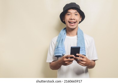 Happy Asian Man Wearing Bright White T-shirt With Black Hat With Blue Towel Playing Phone On Isolated Background