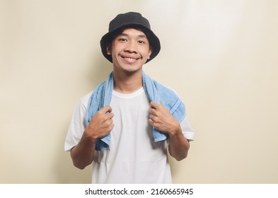 Happy Asian Man Wearing Bright White T-shirt With Black Hat And Carrying Towel On Isolated Background