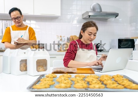 Happy Asian man and woman bakery shop owner working on laptop computer and preparing customer order selling online delivery in kitchen. Small business food and drink occupation entrepreneur concept.