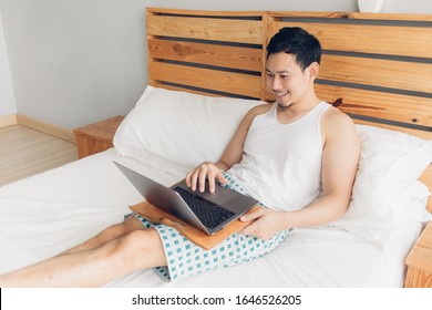 Happy Asian Man Is Working With His Laptop On His Bed. Concept Of Freelancer Successful Lifestyle.