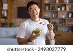 Happy asian mature man holding fork and bowl with fresh vegetable salad, eating healthy lunch after domestic training. Active middle aged male having balanced meal