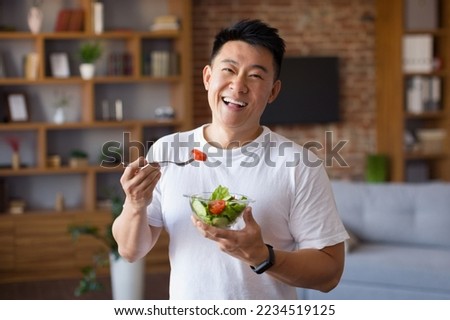 Happy asian mature man holding fork and bowl with fresh vegetable salad, eating healthy lunch after domestic training. Active middle aged male having balanced meal
