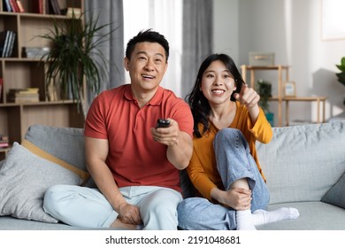 Happy Asian Middle Aged Man And Young Woman In Homewear Sitting On Sofa In Living Room, Watching TV Together At Home, Holding Remote And Pointing At Camera, Copy Space