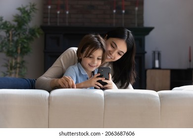 Happy Asian Mom Hugging Joyful Gen Z Kid Excited With Videogame On Smartphone. Younger Mother And Son Boy Playing Virtual Game, Chatting Online, Using App On Mobile Phone, Watching Content On Internet