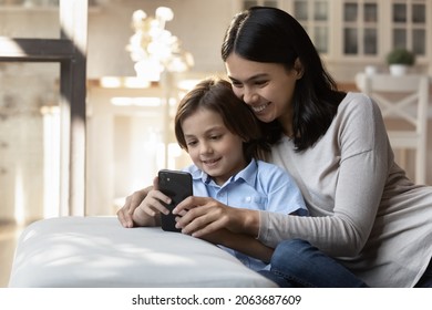 Happy Asian Mom And Little Son Resting On Couch With Cellphone, Chatting Online, Watching Content On Internet, Using App On Mobile Phone. Mother Hugging Kid, Relaxing At Home, Talking On Video Call